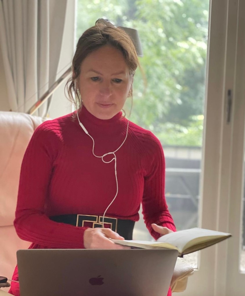 A woman wearing a red sweater and a black belt is sitting at a desk, working on a laptop and holding a notebook, with headphones on. Natural light is coming through a nearby window.